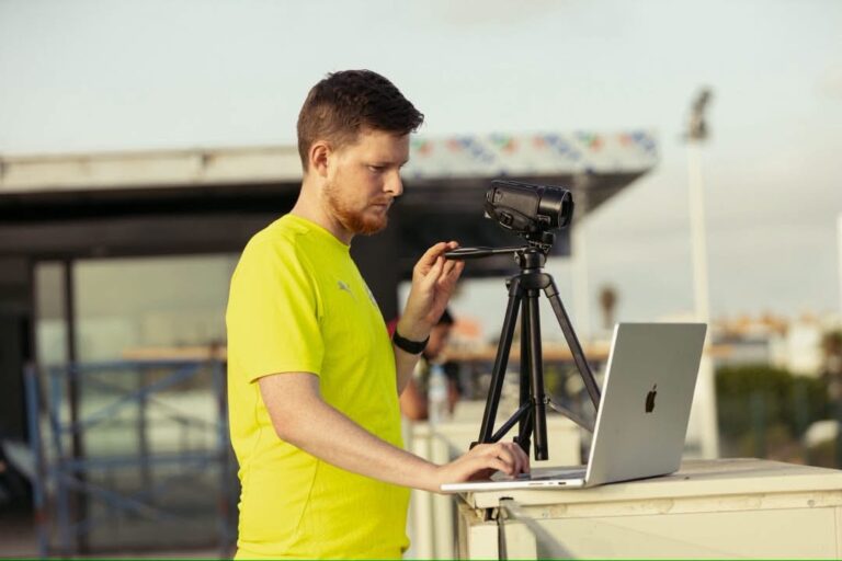 Man in a bright yellow shirt adjusts a camera on a tripod while working on a silver laptop outdoors on a sunny day.