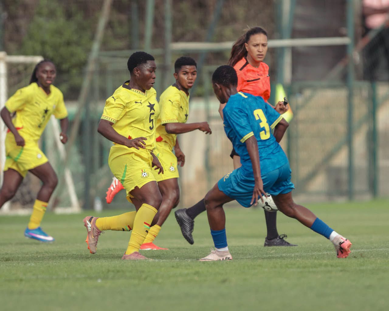 Youth soccer match: players in yellow and blue kits chase the ball on a green field with a referee in the background? (action scene)