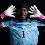 Goalkeeper in a blue patterned jersey with the number 1, wearing white gloves with pink cuffs, smiling against a black background.