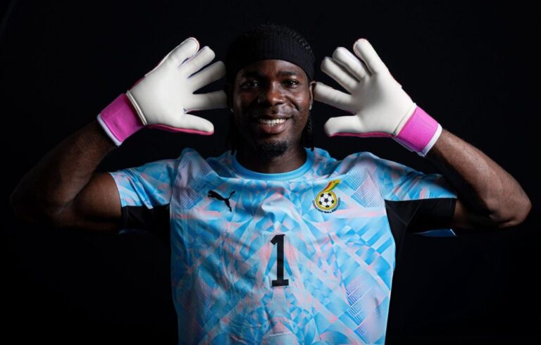 Goalkeeper in a blue patterned jersey with the number 1, wearing white gloves with pink cuffs, smiling against a black background.