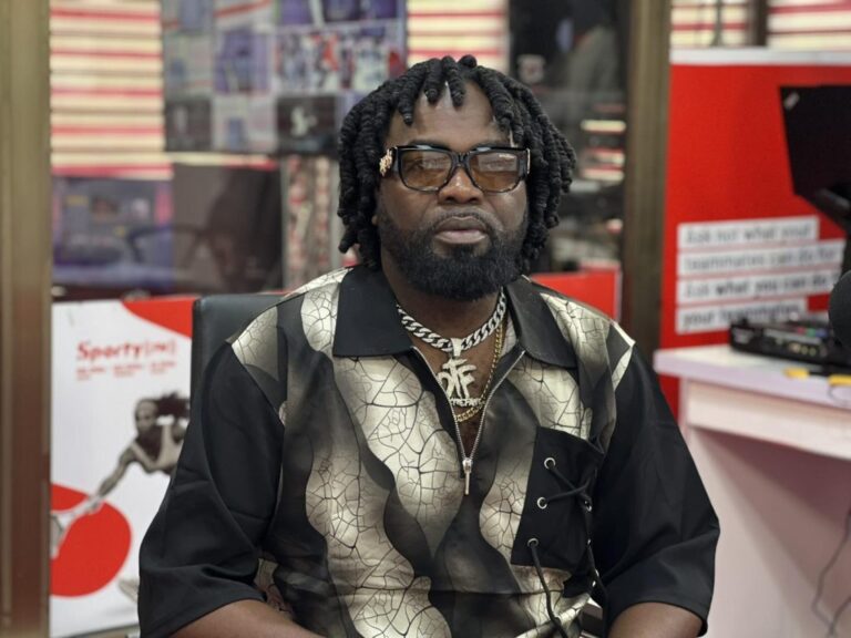 Man with braided hair and sunglasses sits indoors, wearing a patterned black‑beige shirt and gold jewelry.
