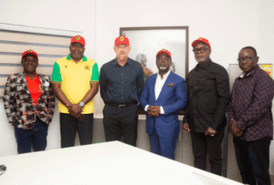 Six people standing indoors in a row, wearing red caps; a woman in a patterned blazer and red top stands with five men in smart-casual outfits posing for a group photo.