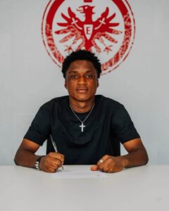 Young man in a black shirt signs a document at a white table, wearing a silver cross necklace, with a red emblem on the wall behind him.