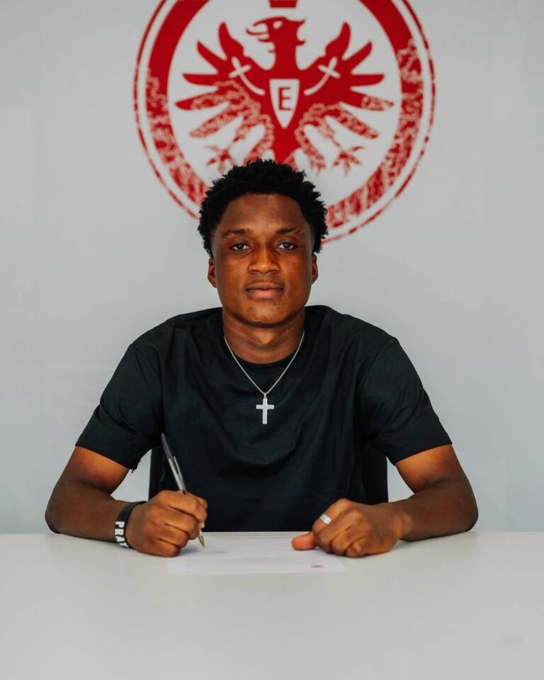 Young man in a black shirt signs a document at a white table, wearing a silver cross necklace, with a red emblem on the wall behind him.