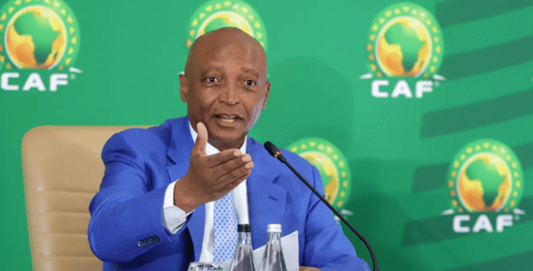 Man in a blue suit gestures while speaking into a microphone at a CAF press conference with a green backdrop and logos behind him.