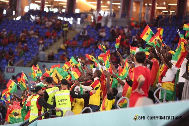 Crowd of football fans in a stadium waving Ghana flags, with security staff in yellow vests along the railing.