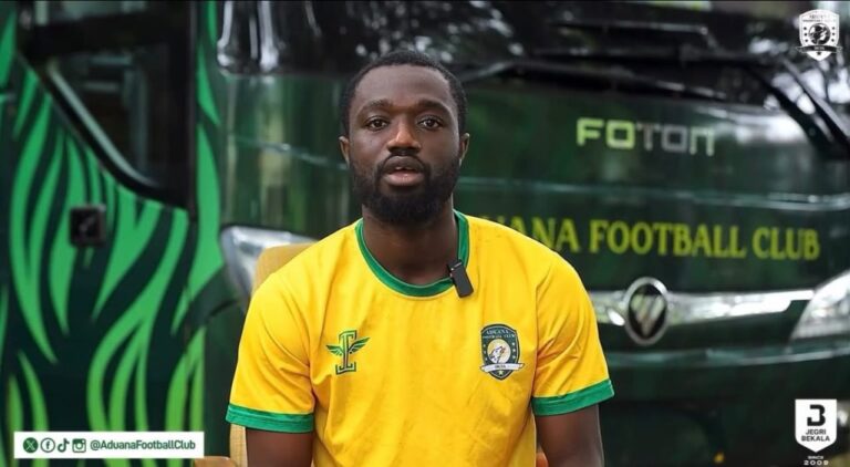 Portrait of a man in a yellow football jersey with green trim, sitting in front of a dark green Aduana Football Club bus with the club crest visible on his chest or nearby.