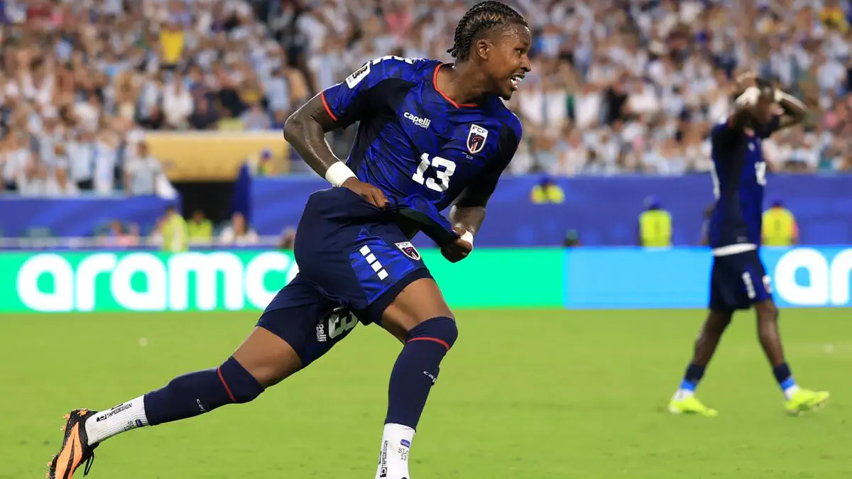 Soccer player in blue kit number 13 sprinting on a green field with cheering fans in the stands behind him.