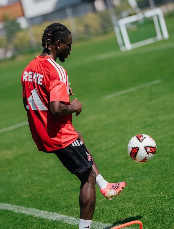 Soccer player in a red shirt and black shorts controlling a soccer ball on a green training field with a goal in the background