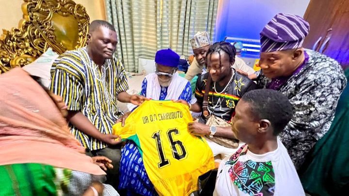 Group of people in colorful attire presenting a yellow sports jersey with the number 16 to a seated recipient in a ceremonial setting.