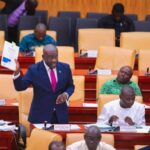 Man in a blue suit and pink tie stands in a parliamentary chamber, holding up a document while colleagues sit and read nearby.