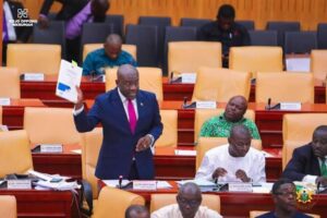 Man in a blue suit and pink tie stands in a parliamentary chamber, holding up a document while colleagues sit and read nearby.