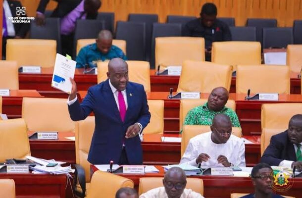 Man in a blue suit and pink tie stands in a parliamentary chamber, holding up a document while colleagues sit and read nearby.
