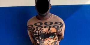 Person with face obscured by a black shape stands against a blue wall wearing an orange shirt with a tiger graphic.