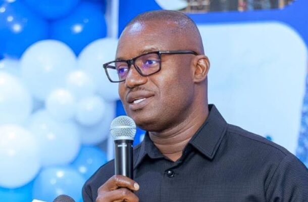 Jinapor | Ghanaguardian.com Dark-skinned man wearing glasses and a black shirt speaks into a handheld microphone at a blue balloon-filled event backdrop.