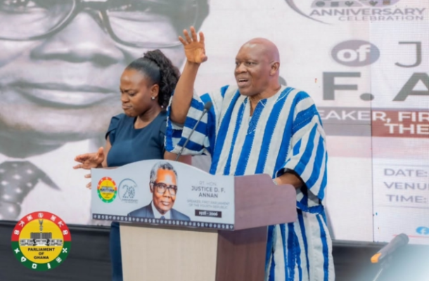 Man in a blue-and-white striped traditional outfit speaks at a podium with one hand raised, flanked by a woman; the lectern features a portrait and the Parliament seal with a background banner.