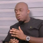 Man with a shaved head wearing a black shirt speaks during an interview, gesturing with hands, seated against a gray paneled wall.