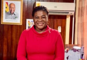 Smiling woman in a red long-sleeve top standing in a wood-paneled room with a framed portrait on the wall.