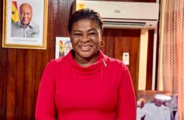 MMMMDDD-1 | Ghanaguardian.com Smiling woman in a red long-sleeve top standing in a wood-paneled room with a framed portrait on the wall.