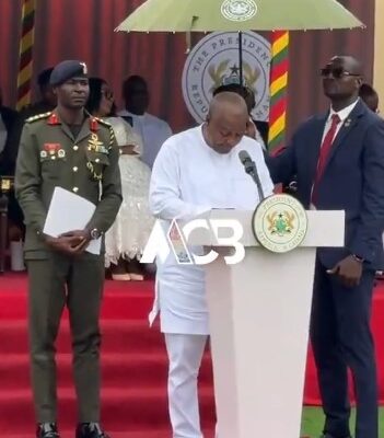 Man in white traditional attire speaks at a podium during a formal ceremony, flanked by a military officer and a suited aide on a red-carpeted stage with a seal in the background.