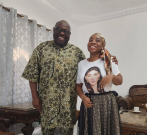 Two adults standing together indoors, smiling at the camera; man in a patterned traditional outfit and woman in a white graphic t-shirt with a scarf and printed skirt.