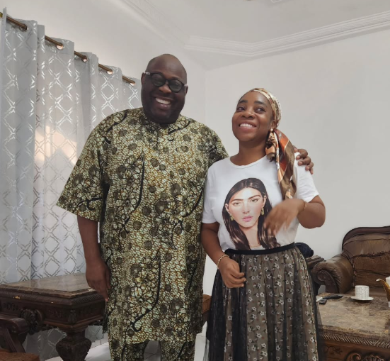 Two adults standing together indoors, smiling at the camera; man in a patterned traditional outfit and woman in a white graphic t-shirt with a scarf and printed skirt.