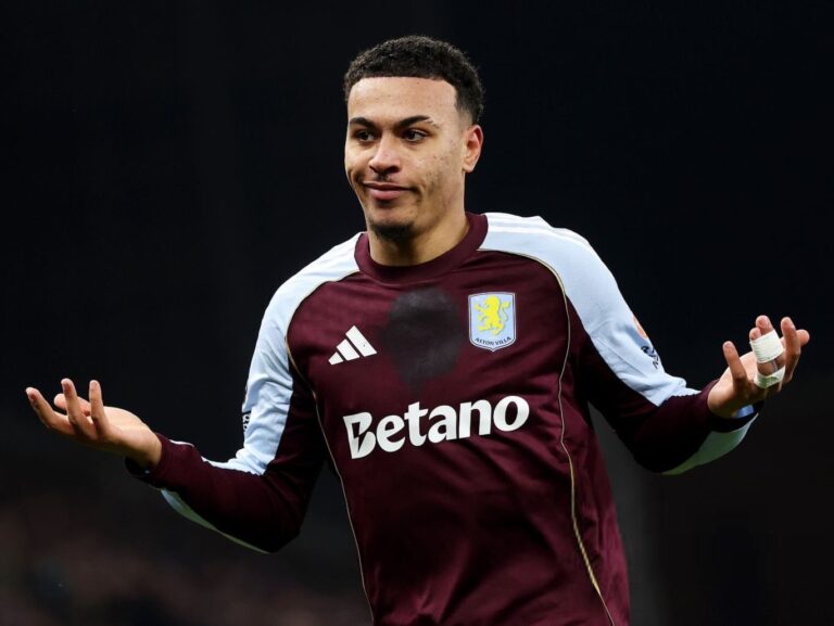 Aston Villa footballer in maroon kit with light-blue sleeves, arms raised in a shrug on the pitch.