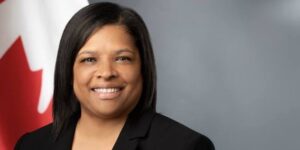 Portrait of a smiling woman in a black blazer with a Canadian flag visible behind her.