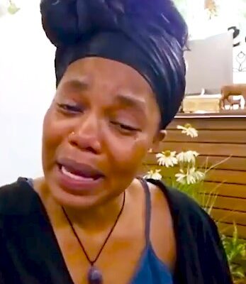 Woman in a black headwrap and blue top crying, tears on her cheeks, appearing distressed in a garden setting with flowers in the background.