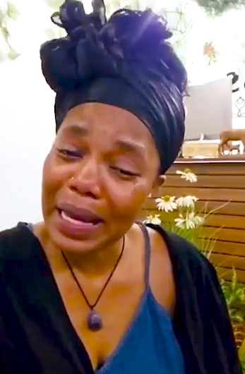 Woman in a black headwrap and blue top crying, tears on her cheeks, appearing distressed in a garden setting with flowers in the background.