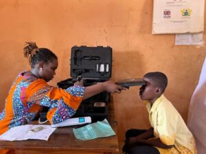A healthcare worker in a bright patterned dress uses a portable medical device to examine a young boy seated at a table with medical equipment nearby.