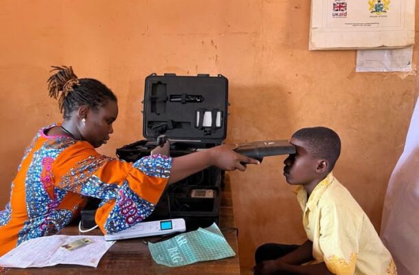 A healthcare worker in a bright patterned dress uses a portable medical device to examine a young boy seated at a table with medical equipment nearby.