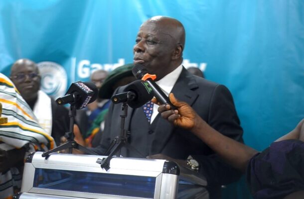Man in a dark suit speaks at a podium with several microphones during a press briefing, with a striped-robed ceremonial bearer on the left and a shirtless man with a microphone on the right against a blue backdrop.