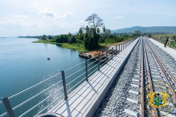 Railroad tracks run along a bridge over a calm river, with a railing on the side and trees in the distance.