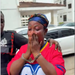 Woman in a red shirt and blue headscarf stands outside, hands pressed together in prayer, appearing distressed while a police officer supports her shoulder.