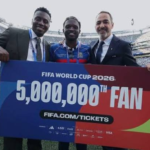 Three men pose with a large colorful banner that reads 'FIFA World Cup 2026 5,000,000th Fan' at a stadium, celebrating the milestone together.