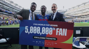 Three men pose with a large colorful banner that reads 'FIFA World Cup 2026 5,000,000th Fan' at a stadium, celebrating the milestone together.