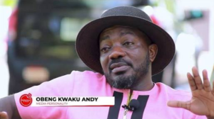 On-screen lower third reads 'Obeng Kwaku Andy, Media Personality'; portrait of a man in a pink shirt and wide-brimmed hat speaking on camera with a microphone clipped to his shirt.