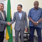 Three men in business attire stand side by side; the man on the left and the man in the center shake hands, with a green-yellow flag behind them.