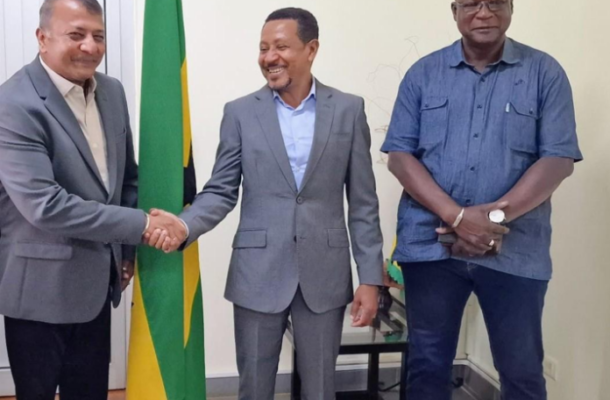 Three men in business attire stand side by side; the man on the left and the man in the center shake hands, with a green-yellow flag behind them.