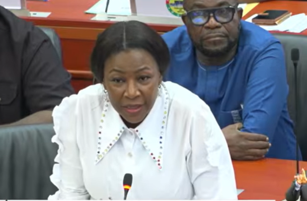 Woman in a white blouse speaks into a microphone at a meeting, with two men seated behind her at a curved conference table (one in blue, one in black).
