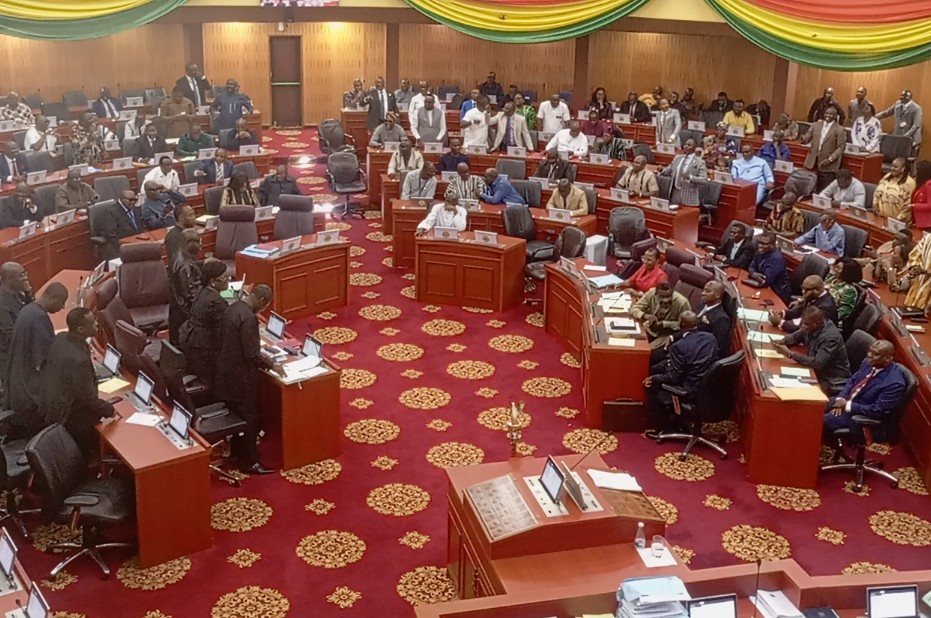 Lawmakers seated around curved wooden desks in a large parliamentary chamber during a session, with many people standing along the walls and a red patterned carpet under bright drapes.