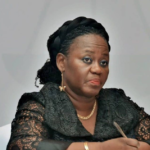 African American woman seated at a table in a black lace outfit, holding a pen and looking thoughtful during a formal meeting.