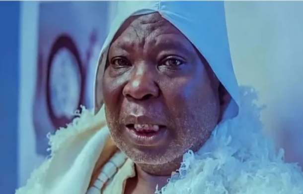 Close-up of a man wearing a white headscarf and robe under blue lighting, looking at the camera with a slight smile.