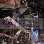 Charred debris and collapsed interior after a fire, with twisted metal and hanging wires among burned furniture pieces.