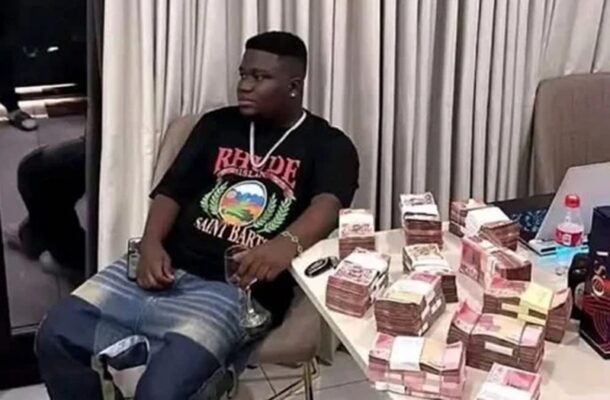WhatsApp-Image-2025-12-16-at-135839 | Ghanaguardian.com Man in a black graphic t-shirt sits in a chair beside a table piled with bundles of cash, holding a glass and looking to the side.