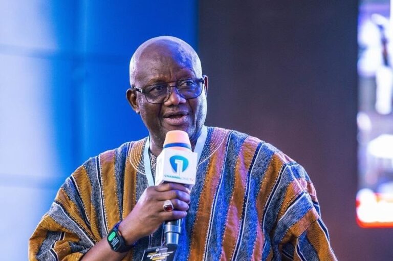 African man wearing glasses in a striped traditional outfit, holding an orange microphone with Channel One TV logo, speaking on stage with a blue backdrop