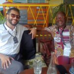 Two men sit on a bench at a cafe, smiling at the camera; one in a white shirt with sunglasses, the other in a colorful patterned shirt with red glasses.