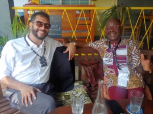 Two men sit on a bench at a cafe, smiling at the camera; one in a white shirt with sunglasses, the other in a colorful patterned shirt with red glasses.