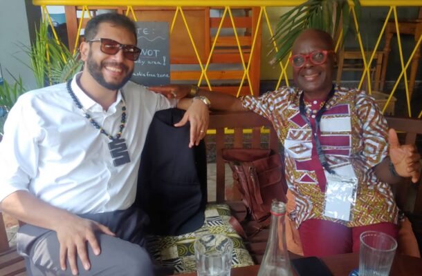 Two men sit on a bench at a cafe, smiling at the camera; one in a white shirt with sunglasses, the other in a colorful patterned shirt with red glasses.